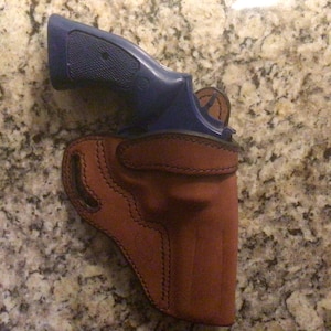Puede incluir: Una funda de cuero marr&oacute;n con un rev&oacute;lver azul en su interior.