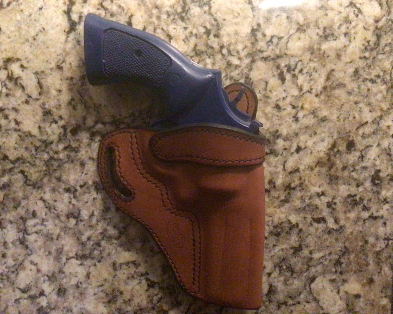 Puede incluir: Una funda de cuero marr&oacute;n con un rev&oacute;lver azul en su interior.