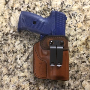 May include: A blue handgun is partially inserted into a brown leather holster. The holster has black metal clips and is designed for concealed carry. The image is taken from a top-down perspective.