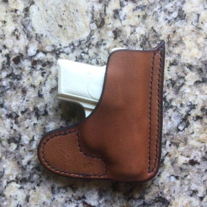 May include: A brown leather holster with a white gun inside. The holster has a stitched design and a brand name on the bottom.