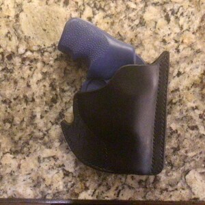 May include: A black leather holster with a stitched edge, holding a blue handgun. The holster is designed to securely carry a firearm. The background is a speckled, light brown surface.