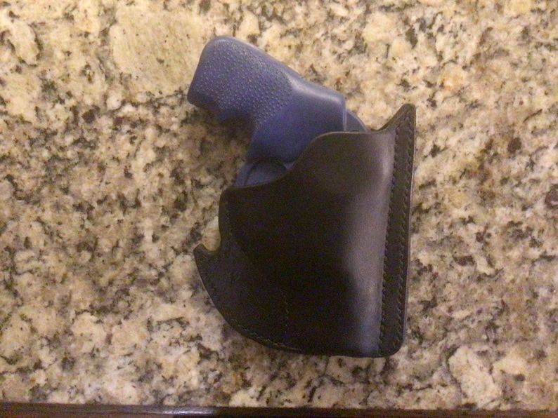 May include: A black leather holster with a stitched edge, holding a blue handgun. The holster is designed to securely carry a firearm. The background is a speckled, light brown surface.