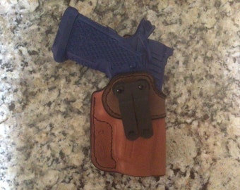 Op de afbeelding: Een bruine leren holster met een zwarte clip voor een blauw pistool.