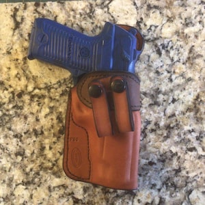 Pode incluir: Um coldre de couro marrom com uma pistola azul dentro. O coldre tem um fecho de press&atilde;o e um passador de cinto. O coldre &eacute; feito por "PRD".