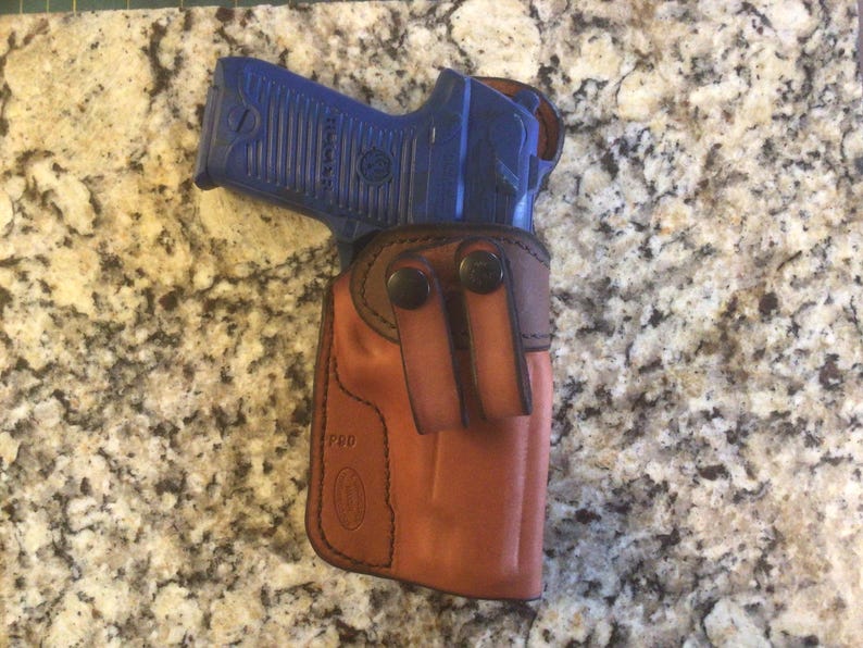 Pode incluir: Um coldre de couro marrom com uma pistola azul dentro. O coldre tem um fecho de press&atilde;o e um passador de cinto. O coldre &eacute; feito por "PRD".