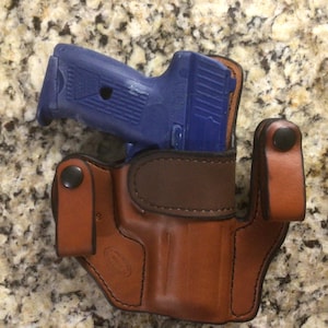 May include: A blue handgun rests in a brown leather holster. The holster has a dark brown interior and is secured with black snaps. The image is taken from a top-down perspective, showcasing the firearm and its carrying case.