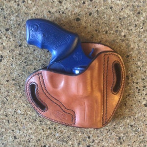 May include: A brown leather holster with a blue revolver inside. The holster has a stitched design and a maker's mark.