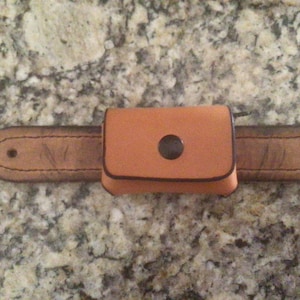 May include: A brown leather belt with a small, rectangular pouch attached. The pouch has a brown leather flap with a metal snap closure. The belt has two metal eyelets on each side.