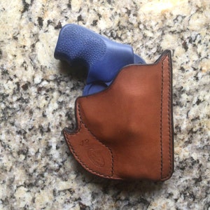 May include: A brown leather holster with black stitching, holding a blue handgun. The holster has a curved design and a stamped logo near the bottom. The background is a speckled, light-colored surface.
