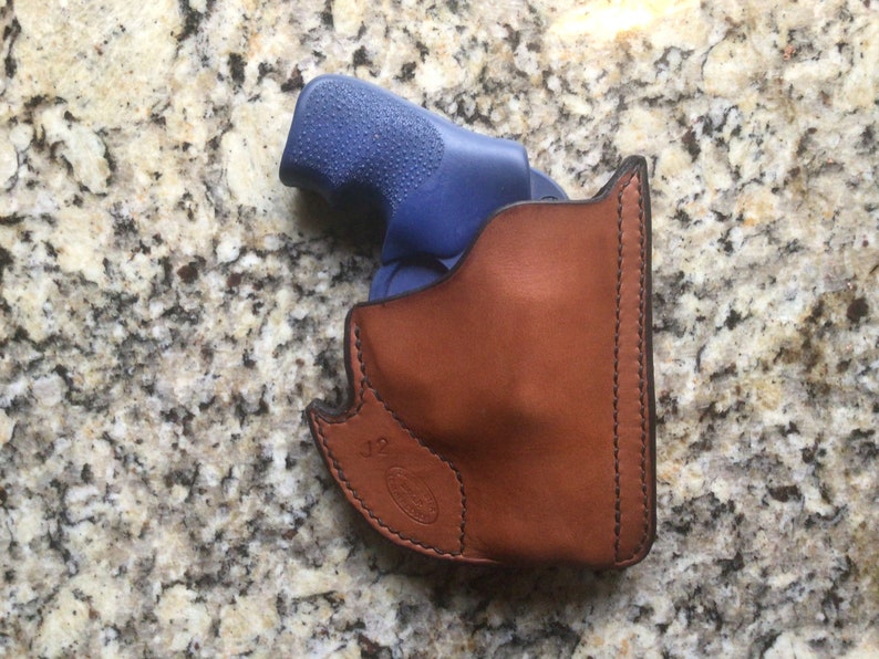 May include: A brown leather holster with black stitching, holding a blue handgun. The holster has a curved design and a stamped logo near the bottom. The background is a speckled, light-colored surface.