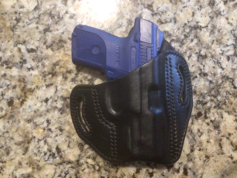 May include: A black leather holster with a blue handgun inside. The holster has stitching around the edges and a belt loop.