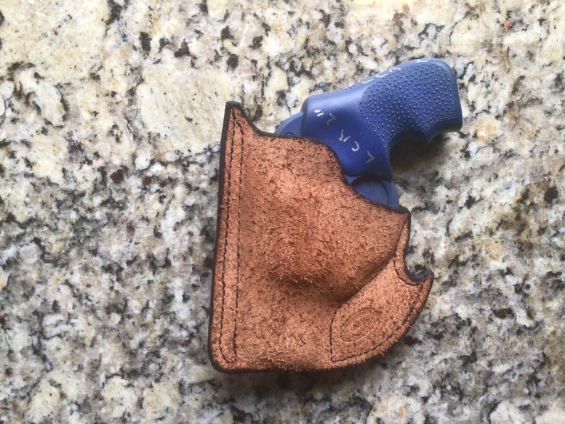May include: A brown leather holster with a blue handgun inside. The holster is made of textured leather and has black stitching around the edges. The handgun has a blue grip. The image is taken from a top-down perspective.
