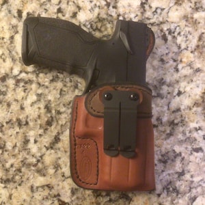 May include: A brown leather holster with a black clip for a handgun. The holster has a brown leather flap with a stitched design and the word "FREE" embossed on it.