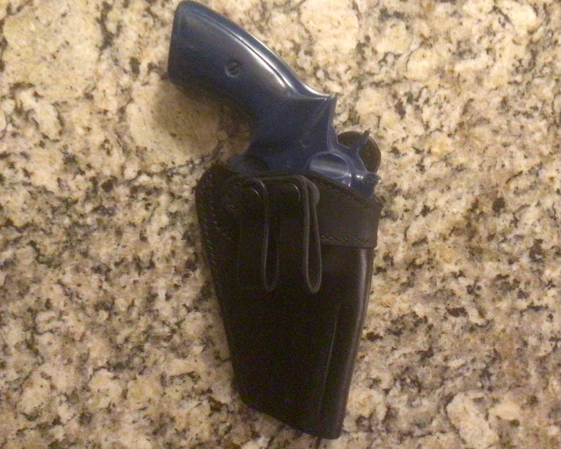 Puede incluir: Un rev&oacute;lver azul en una funda de cuero negro. La funda tiene un bucle para el cintur&oacute;n y un cierre a presi&oacute;n.