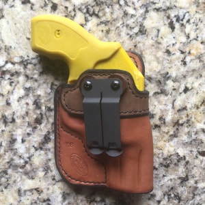 May include: A yellow handgun in a brown leather holster with a black clip. The holster has a brown leather flap and a black metal clip.