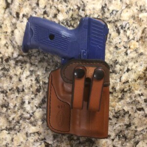 May include: A blue handgun rests in a brown leather holster. The holster has two belt loops and black snap closures. The background is a speckled, light brown surface.