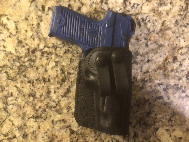 Pode incluir: Um coldre de couro preto com uma pistola azul dentro. O coldre tem um clipe de cinto e um fecho de press&atilde;o.