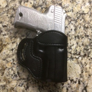 May include: A silver handgun rests partially inside a black leather holster. The holster is designed to fit the gun's shape, with visible stitching and a belt loop. The background is a speckled, light brown surface.