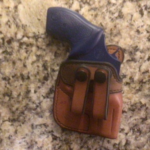 May include: A brown leather holster with a blue handgun inside. The holster has two metal clips on the back.