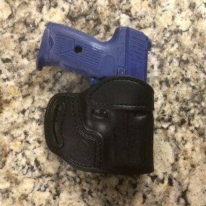 May include: A blue handgun rests atop a black leather holster. The handgun has a textured grip and slide. The holster is made of smooth leather with visible stitching.
