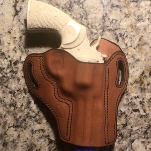 May include: A brown leather holster with a white handgun inside. The holster has a stitched design and a belt loop.