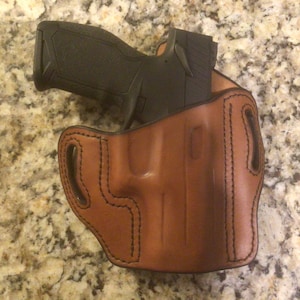 May include: A brown leather holster with a black handgun inside. The holster has stitching around the edges and a cut-out for the trigger guard.