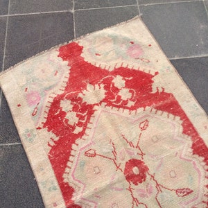 May include: A vintage rug with a cream background and a red floral pattern. The rug has a fringed edge and is made of wool.