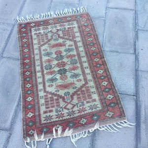 Puede incluir: Una alfombra pequeña y rectangular con fondo crema y un patrón geométrico rojo, verde y azul. La alfombra tiene un borde de flecos.