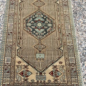 May include: A rectangular area rug featuring a central geometric medallion in shades of teal, dark blue, and deep red against a tan background. The rug is decorated with intricate linear patterns, repeating motifs, and a detailed border design.