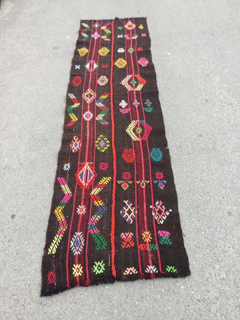 Puede incluir: Una alfombra larga y estrecha con un fondo marr&oacute;n oscuro y patrones geom&eacute;tricos coloridos. La alfombra tiene flecos en ambos extremos.