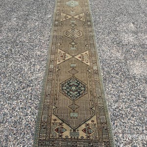 May include: A long, narrow runner rug featuring a beige and tan base with intricate geometric patterns in shades of sage green, dark blue, and deep red. The design includes repeating diamond-shaped medallions and a detailed border pattern.