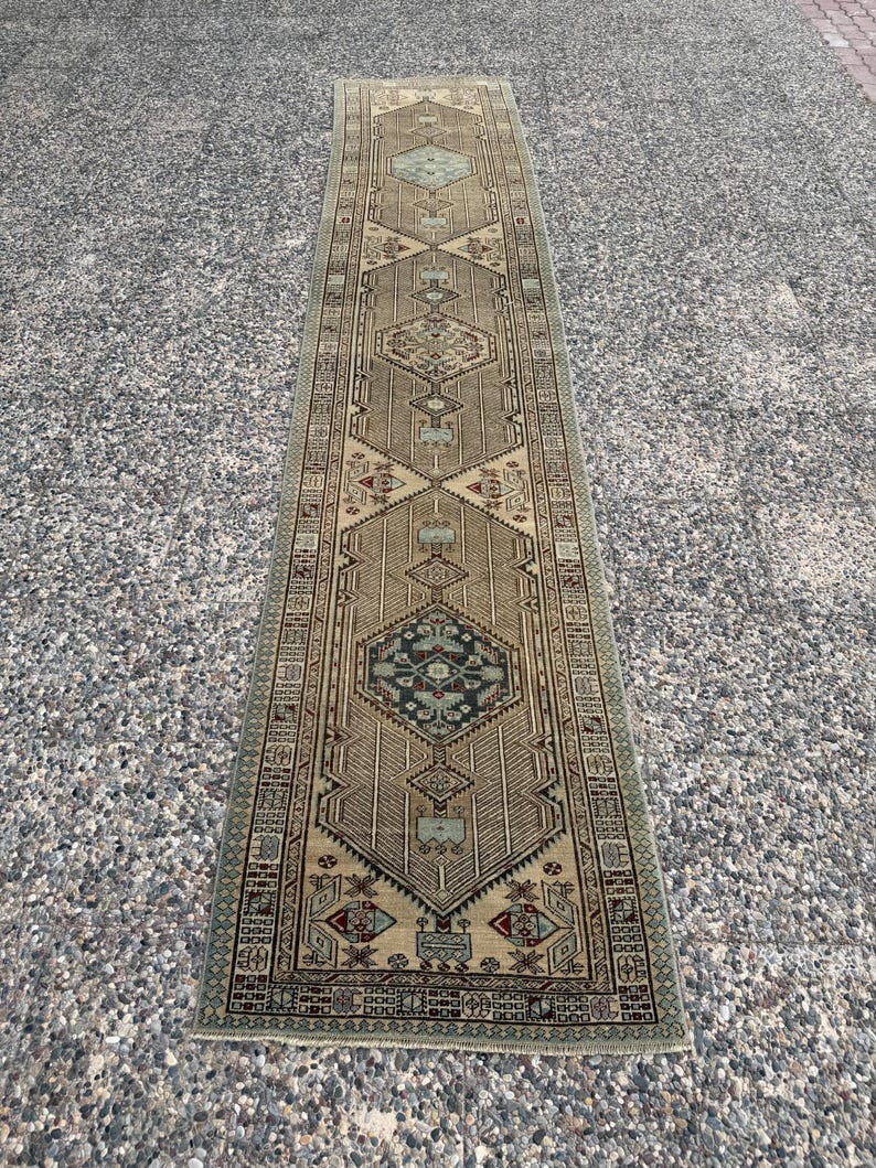 May include: A long, narrow runner rug featuring a beige and tan base with intricate geometric patterns in shades of sage green, dark blue, and deep red. The design includes repeating diamond-shaped medallions and a detailed border pattern.