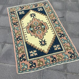 Puede incluir: Una alfombra vintage con un patrón geométrico tradicional en tonos rosa, azul y crema. La alfombra tiene un borde con flecos y está hecha de lana.