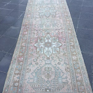 May include: A vintage Turkish runner rug with a light pink background and a geometric pattern in shades of green, brown, and white. The rug has a fringed edge.
