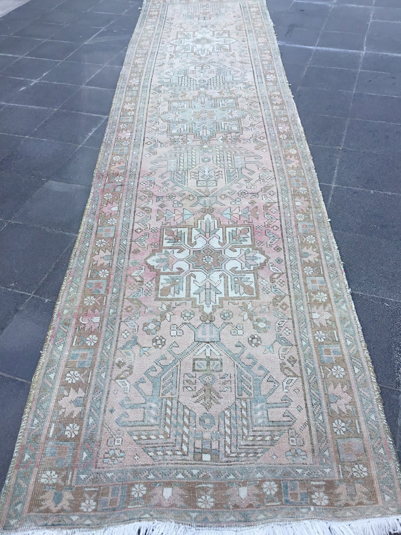 May include: A vintage Turkish runner rug with a light pink background and a geometric pattern in shades of green, brown, and white. The rug has a fringed edge.