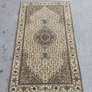 Puede incluir: Una alfombra vintage de color crema con un patrón floral detallado en tonos marrones, verdes y negros. La alfombra tiene un medallón central y un borde con un diseño repetitivo.