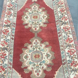 Puede incluir: Una alfombra roja vintage con un centro de color crema y intrincados patrones florales en tonos de azul, verde y rojo. La alfombra tiene un aspecto descolorido y una textura desgastada.