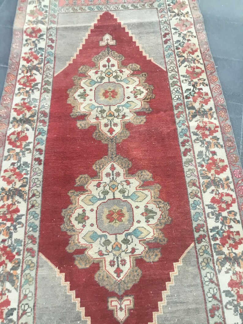 Puede incluir: Una alfombra roja vintage con un centro de color crema y intrincados patrones florales en tonos de azul, verde y rojo. La alfombra tiene un aspecto descolorido y una textura desgastada.