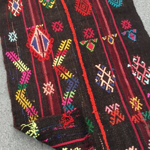 Puede incluir: Una alfombra colorida tejida a mano con intrincados patrones geom&eacute;tricos en tonos de rojo, azul, verde, amarillo y rosa sobre un fondo marr&oacute;n oscuro.