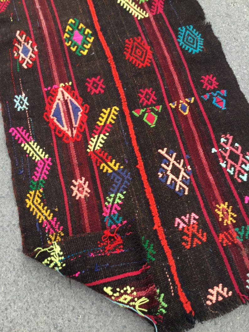 Puede incluir: Una alfombra colorida tejida a mano con intrincados patrones geom&eacute;tricos en tonos de rojo, azul, verde, amarillo y rosa sobre un fondo marr&oacute;n oscuro.