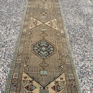 May include: A long, narrow runner rug featuring a tan background with a repeating geometric pattern in shades of dark blue, light blue, and deep red. The design includes central diamond motifs, intricate borders, and symmetrical linear details.