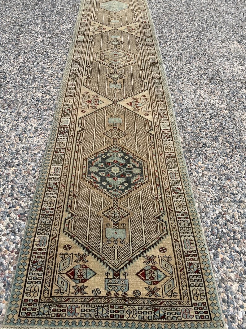 May include: A long, narrow runner rug featuring a tan background with a repeating geometric pattern in shades of dark blue, light blue, and deep red. The design includes central diamond motifs, intricate borders, and symmetrical linear details.
