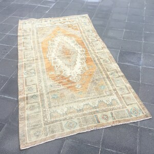 Puede incluir: Una alfombra vintage con fondo beige y un medallón central en tonos naranja y marrón. La alfombra tiene un borde decorativo con patrones geométricos en tonos azul, verde y marrón.