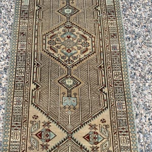 May include: A long rectangular runner rug featuring a beige background with intricate geometric patterns in shades of light blue, dark red, and brown. The design includes a central diamond motif, repeating border elements, and symmetrical linear details.