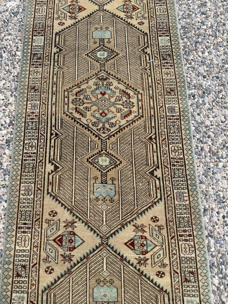 May include: A long rectangular runner rug featuring a beige background with intricate geometric patterns in shades of light blue, dark red, and brown. The design includes a central diamond motif, repeating border elements, and symmetrical linear details.