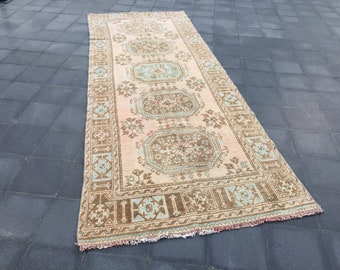 Puede incluir: Una alfombra rectangular larga con fondo rosa claro y un patrón floral marrón y azul. La alfombra tiene un borde de flecos.