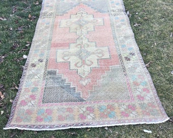May include: A vintage Turkish rug with a faded pink, green, and blue pattern. The rug has a fringed edge and is laid out on a grassy surface.