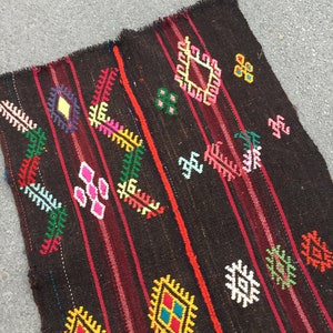 Puede incluir: Una alfombra tejida de color marr&oacute;n oscuro con patrones geom&eacute;tricos coloridos. La alfombra tiene un borde rayado rojo y negro y presenta una variedad de formas de diamantes y flores.