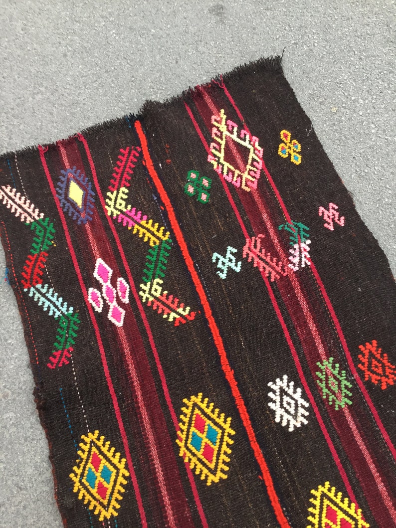 Puede incluir: Una alfombra tejida de color marr&oacute;n oscuro con patrones geom&eacute;tricos coloridos. La alfombra tiene un borde rayado rojo y negro y presenta una variedad de formas de diamantes y flores.