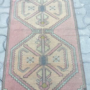 Peut inclure: Un tapis rectangulaire de style vintage, aux tons rose et beige. Il pr&eacute;sente un motif g&eacute;om&eacute;trique central avec des formes r&eacute;p&eacute;t&eacute;es et une bordure en zigzag. Le tapis est probablement en mati&egrave;re tiss&eacute;e.